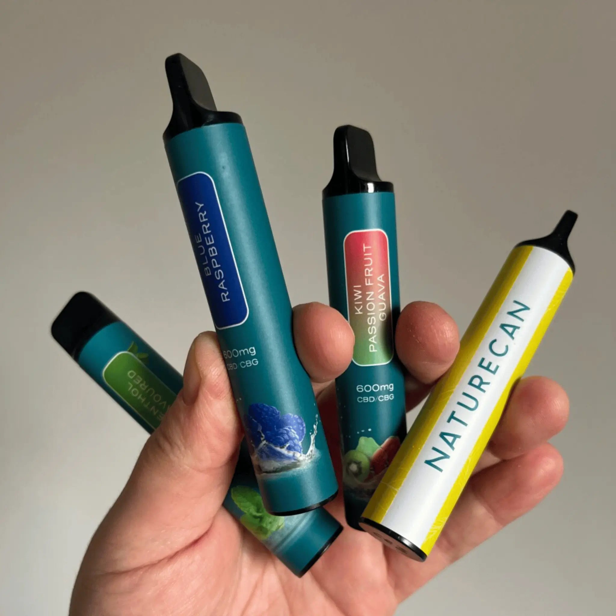 Hand holding colorful CBD vape pens labeled with flavors like Blue Raspberry and Kiwi Passion Fruit.