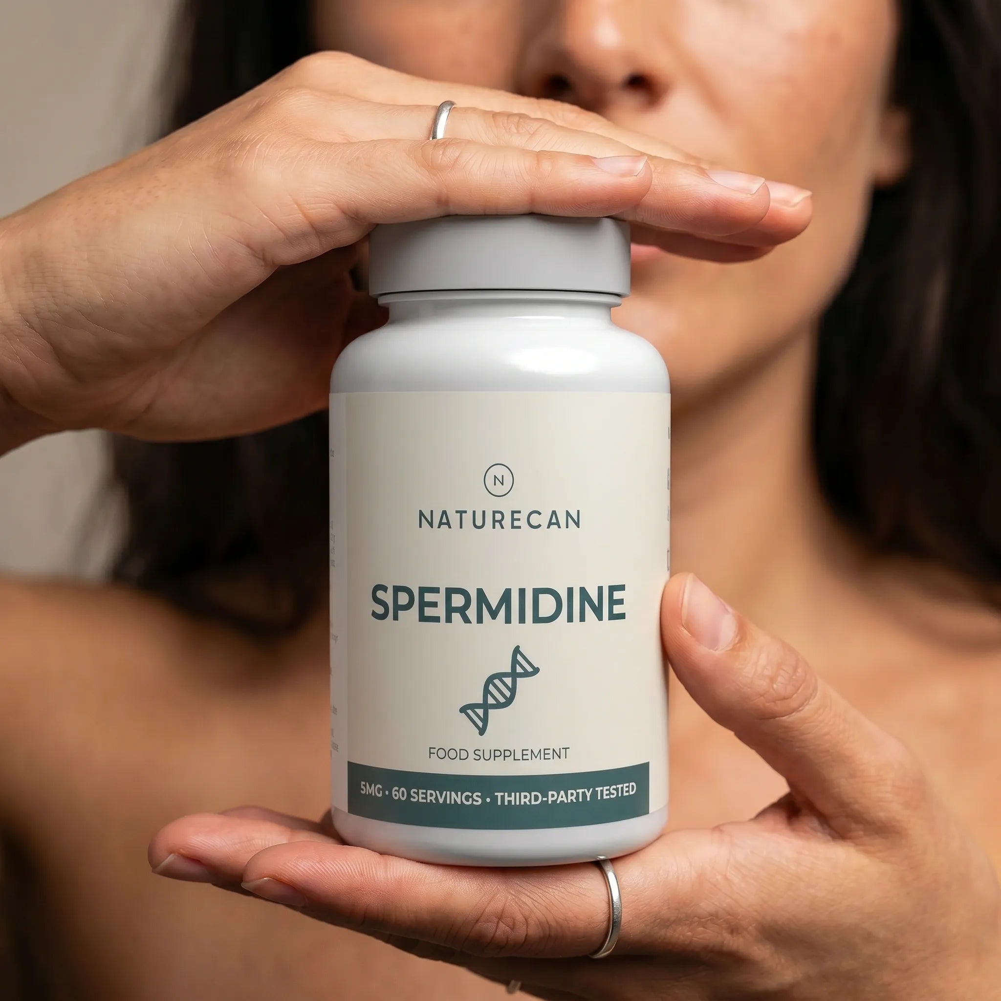 A close-up of a person holding a white bottle of Naturecan Spermidine food supplement, with their hands framing the bottle and a blurred face in the background.