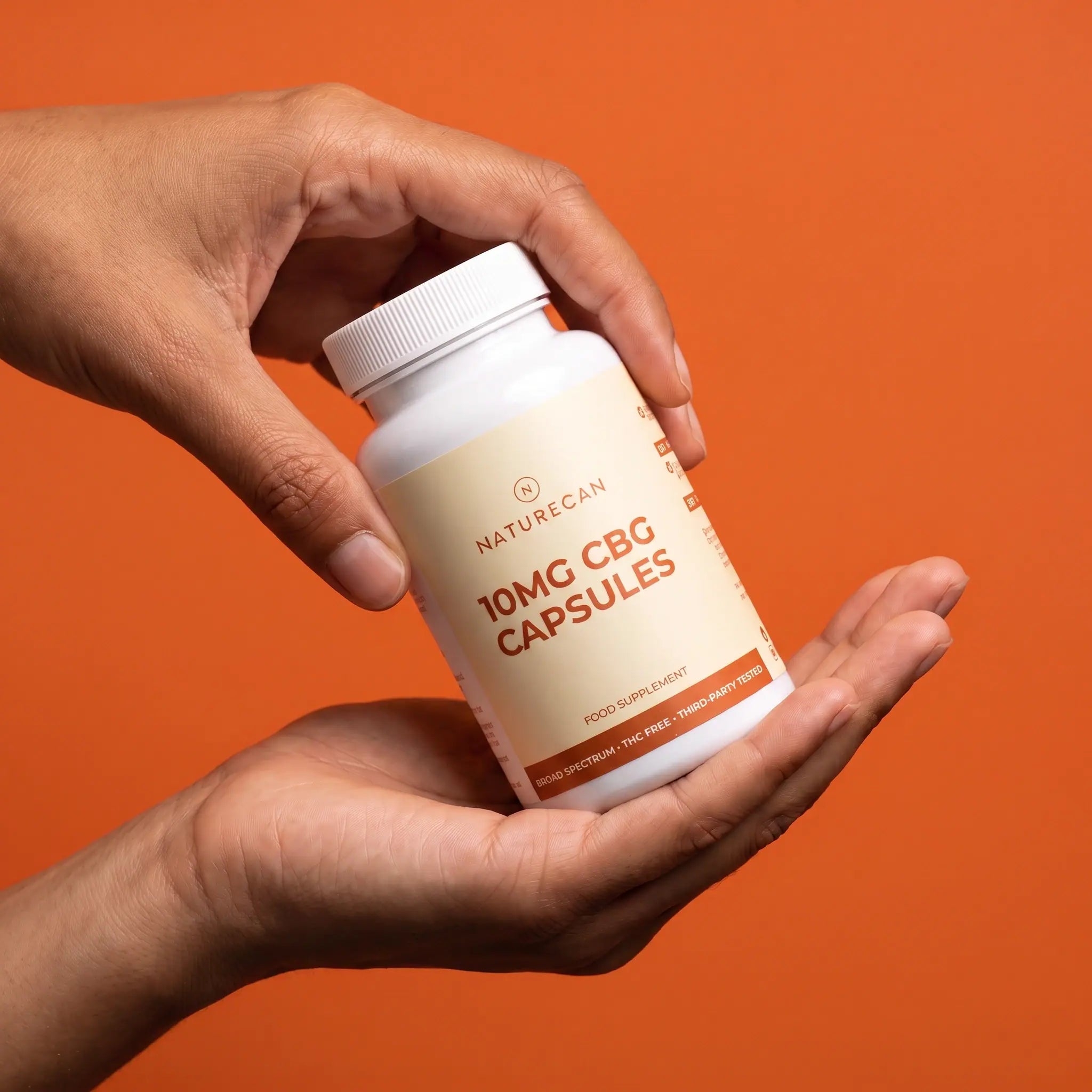 A person's hands holding a white bottle of NatureCan 10mg CBG capsules against a solid orange background.