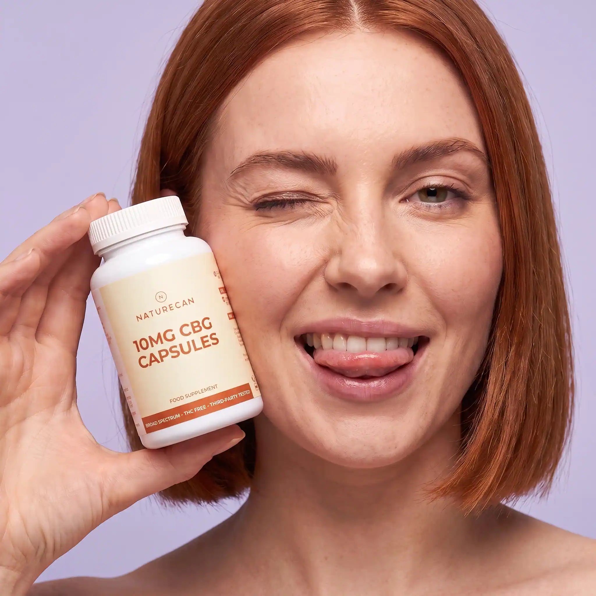 A smiling woman with red hair holds a white bottle of Naturecan 10mg CBG capsules next to her face against a light purple background.