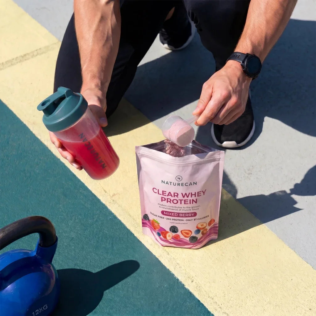 Person holding a protein shake and Naturecan Clear Whey Protein package on a colorful outdoor floor.