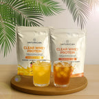 Naturecan Clear Whey Protein packages with two glasses of flavored tea on a wooden board against a leafy green background.