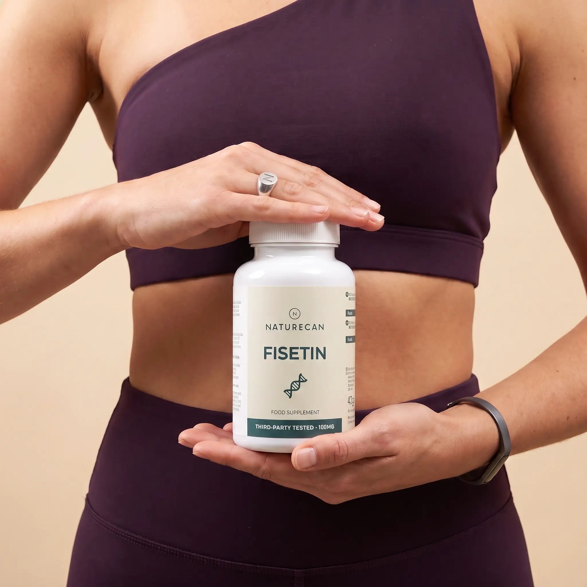 A woman in a dark purple sports bra and leggings holds a white bottle of Naturecan Fisetin dietary supplement against a solid beige background.