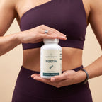 A woman in a dark purple sports bra and leggings holds a white bottle of Naturecan Fisetin dietary supplement against a solid beige background.