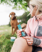 Woman in pink jacket holding Naturecan product, with a brown dog in the background.