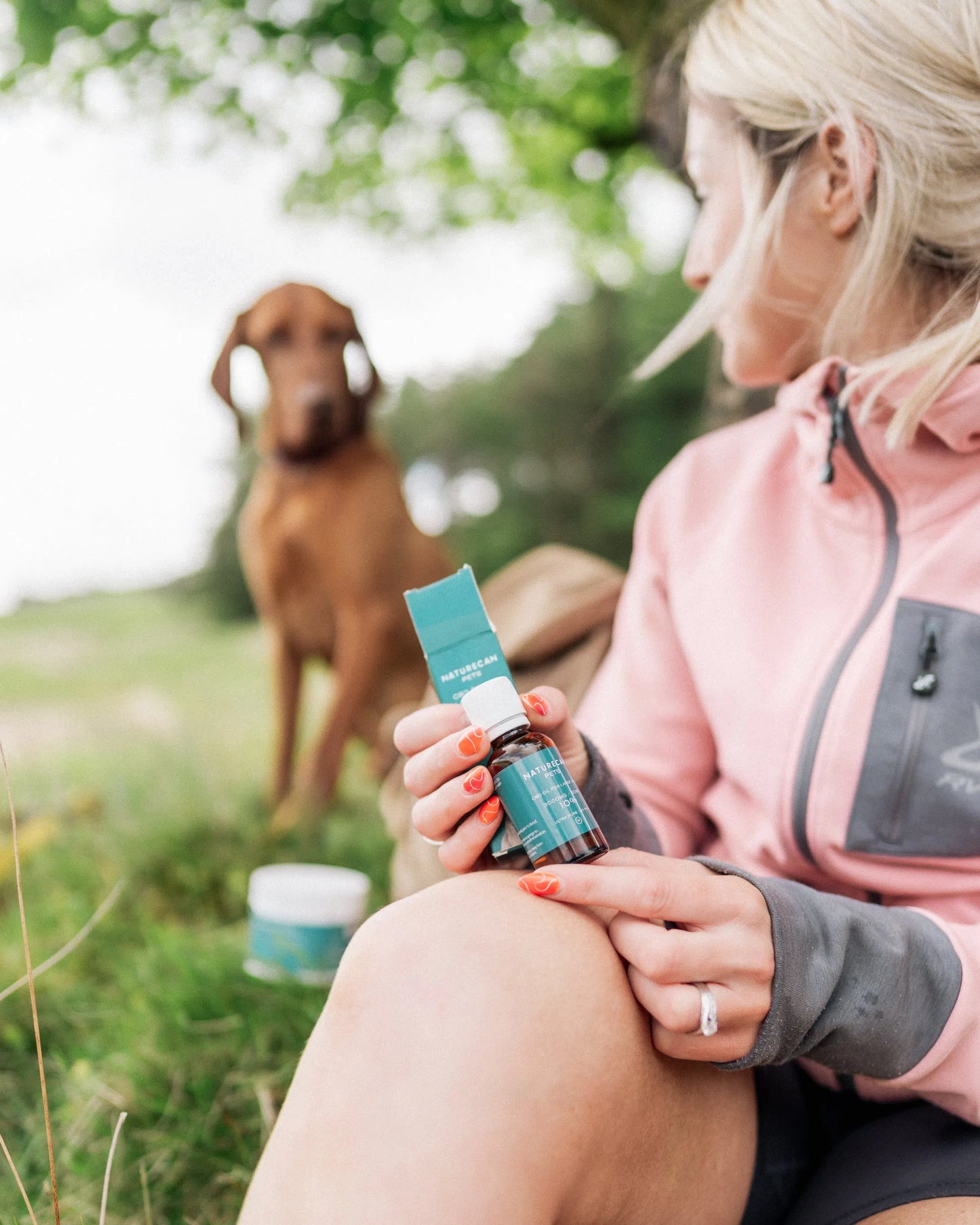 Woman in pink jacket holding Naturecan product, with a brown dog in the background.