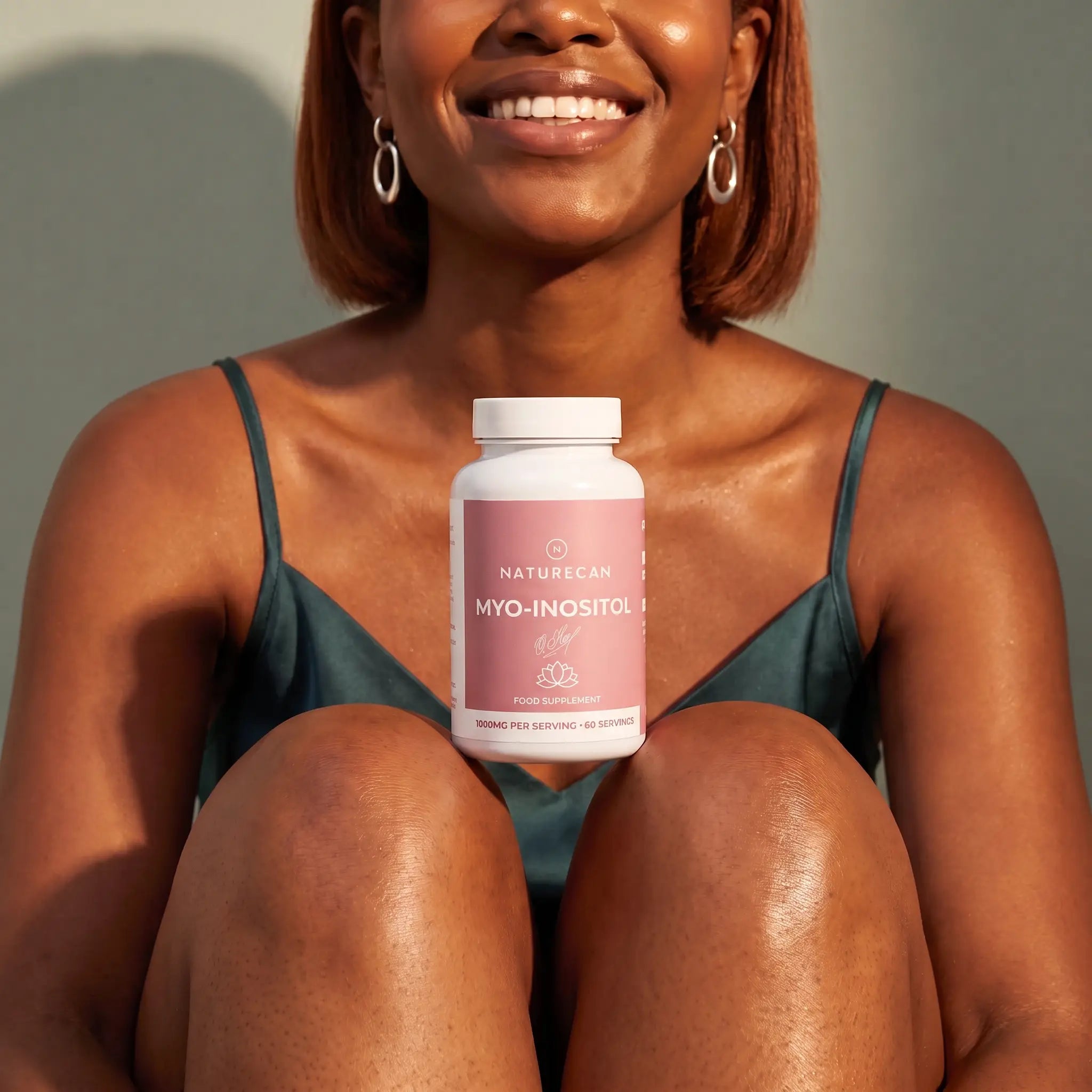 A smiling woman in a teal satin camisole sits with her knees drawn up, holding a white bottle of NatureCan Myo-Inositol supplements. The background is a clean, neutral gray wall with warm sunlight casting soft shadows.