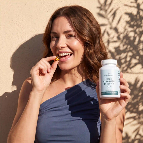 A smiling woman in a navy one-shoulder top holds a white bottle of Merivita High Strength Omega 3 while eating a gold capsule in her mouth, set against a warm beige wall with leaf shadows.