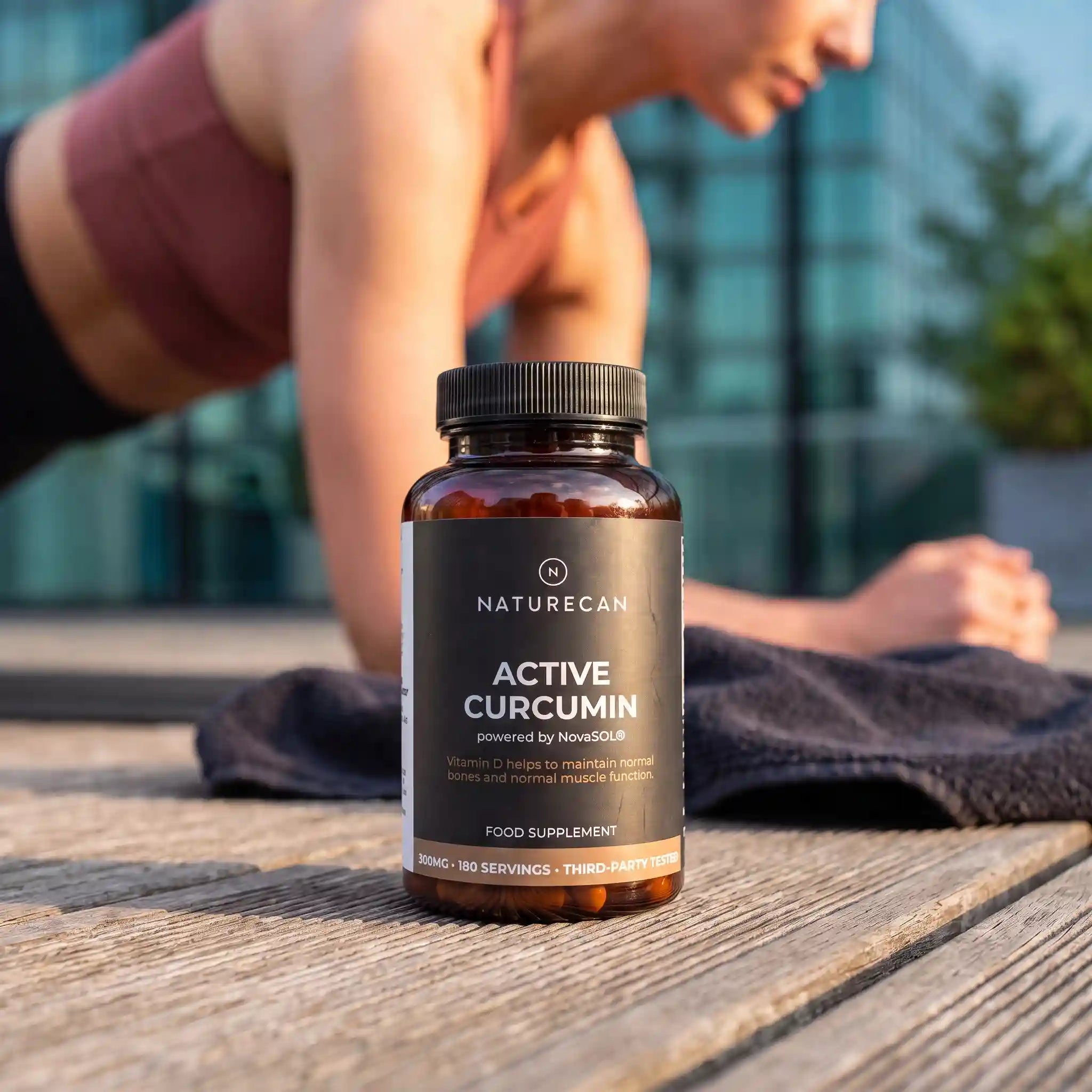 A bottle of Naturecan Active Curcumin food supplement sits on a wooden deck in the foreground, with a woman in athletic wear blurred in the background near a glass building.